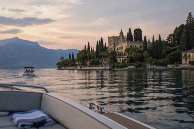 A small private boat approaches Isola del Garda at sunset, the island's gardens and pale villa glowing against dark hills while the water carries long streaks of gold.
