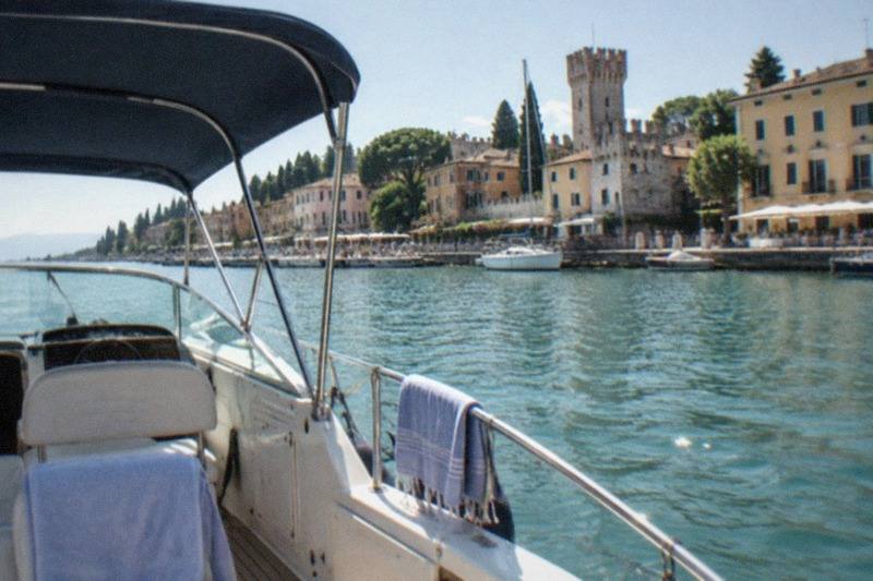 From a white motorboat with blue canopy and towels on empty seats, looking across water at a Lake Garda lakeside town with a stone tower and buildings.