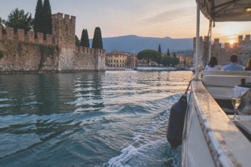 From a boat at sunset, a stone castle wall and cypresses line the left shore, with seated passengers, a wine glass, and distant hills ahead, on Lake Garda.