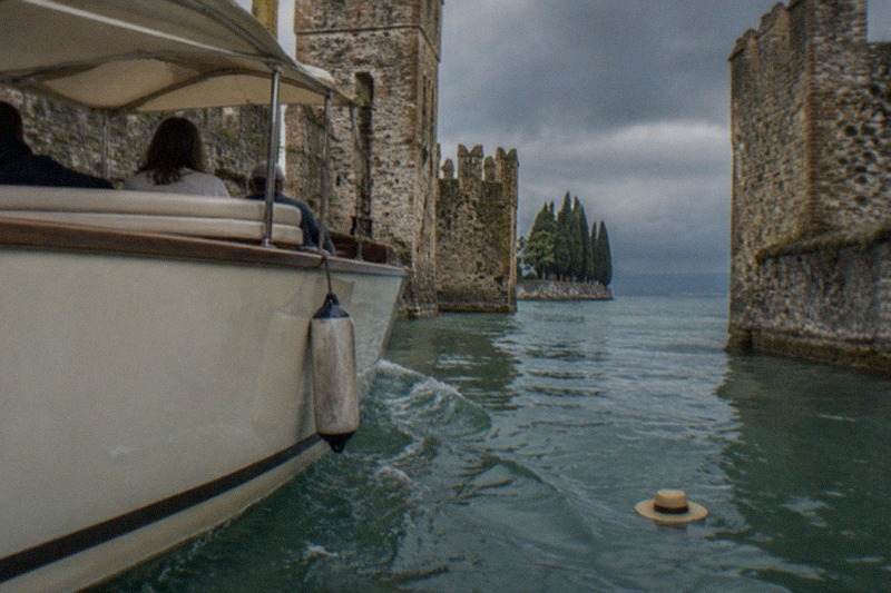 A white motorboat with passengers passes between stone castle walls on water, a straw hat floating in the wake under cloudy skies, on Lake Garda.