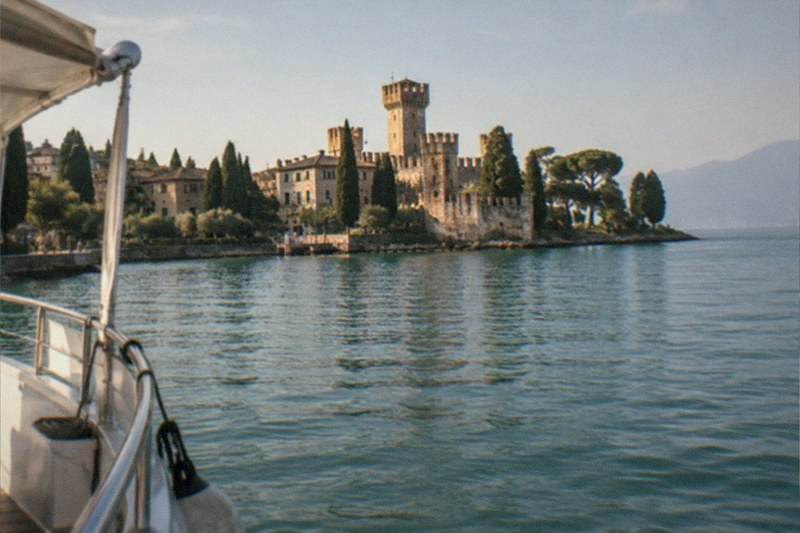 From a boat's bow, a stone castle with towers and cypress trees sits on a lakeshore under a pale sky, on Lake Garda.