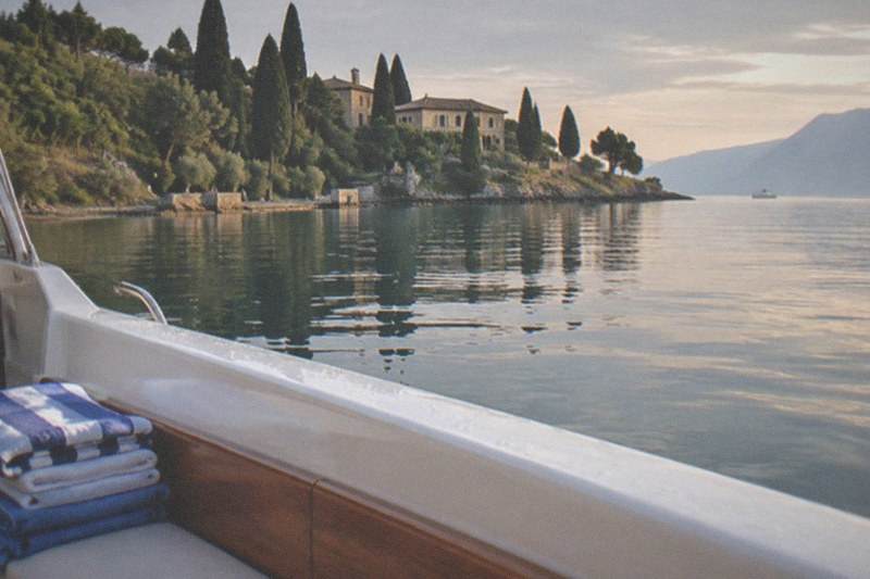 From a boat's white gunwale with stacked towels, a Lake Garda lakeside villa among cypress trees is reflected in calm water with mountains beyond.