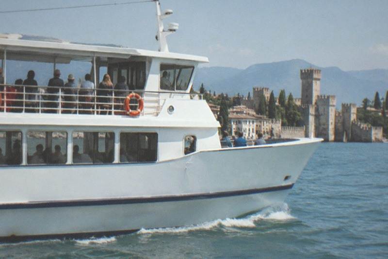A white passenger boat with people on the upper deck moves on blue water toward a Lake Garda lakeside castle with towers and mountains behind.