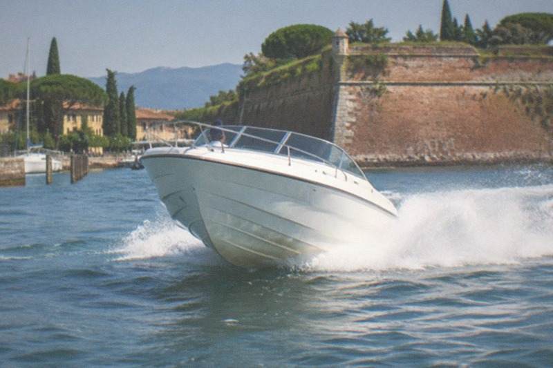 A white motorboat speeds toward the camera throwing bow spray, with a stone fortress wall, trees and shoreline buildings behind it, on Lake Garda.