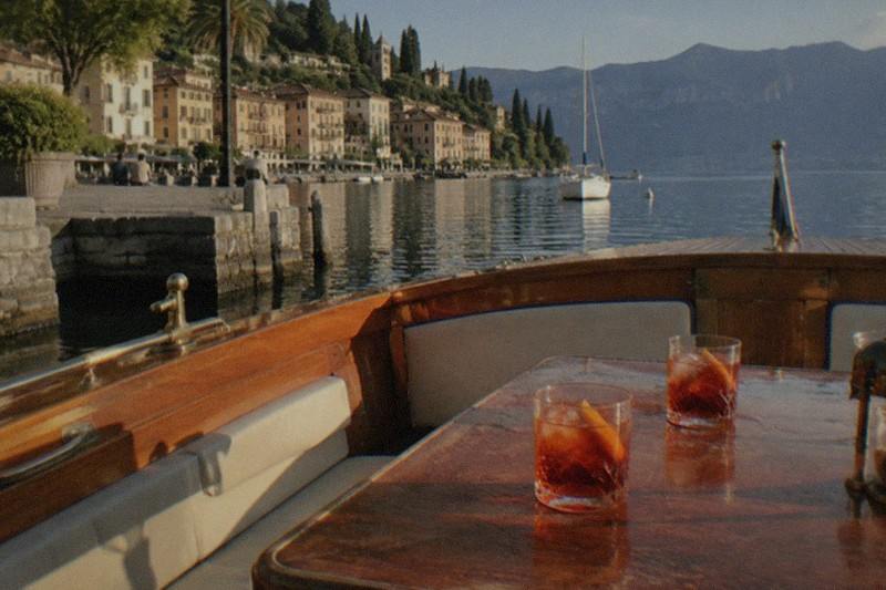 From a wooden boat with brass fittings and two amber drinks on a table, looking toward a Lake Garda lakeside town, trees, a sailboat and mountains.