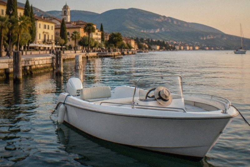 A white motorboat with a hat on the windshield sits moored at a stone quay, Lake Garda lakeside buildings and mountains behind in warm evening light.