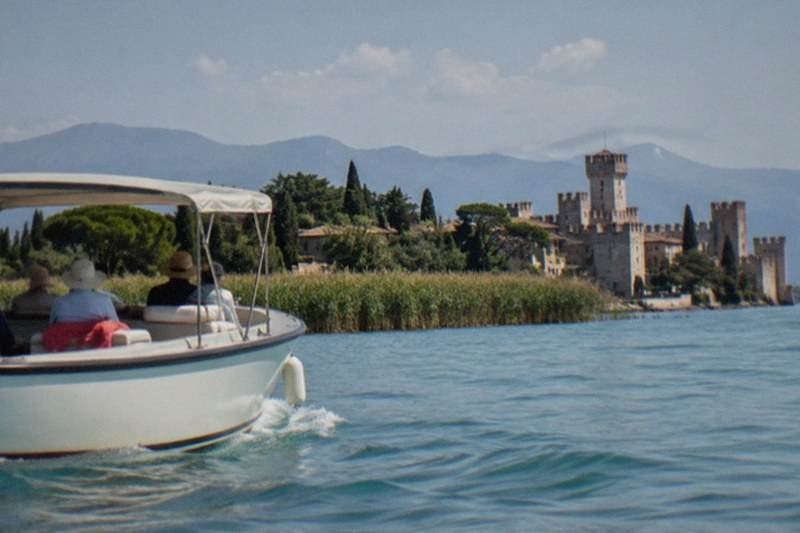 Small white motorboat with passengers on Lake Garda, heading toward a lakeside castle with towers, trees, reeds and mountains beyond.