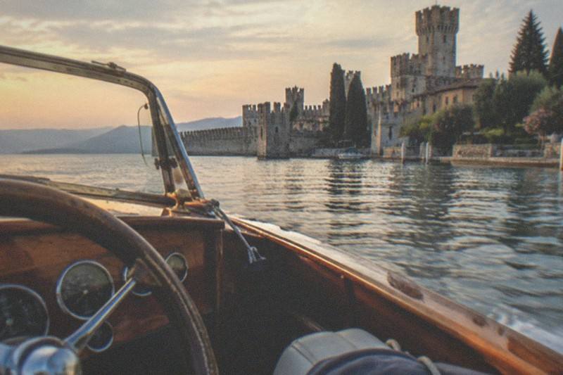 From a wooden motorboat helm, the empty wheel and dashboard face a Lake Garda lakeside castle and trees across calm water at sunset.