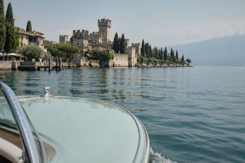 From a pale boat bow, a Lake Garda lakeside castle with towers and cypress trees lines the shore under a clear sky, mountains beyond.