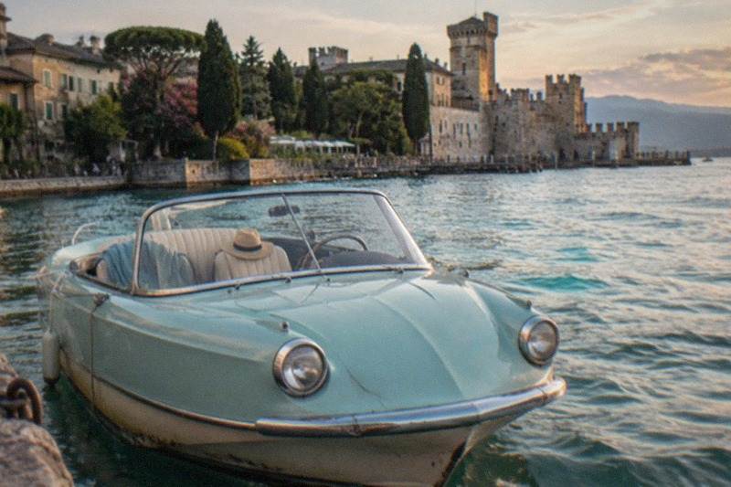 A pale mint vintage car-shaped boat with round headlights floats on still water before a stone castle, trees and Lake Garda lakeside houses at dusk.