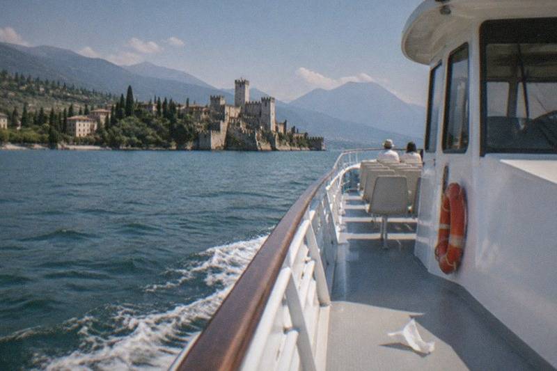From a boat deck with a life ring and two seated passengers, looking across the wake toward a Lake Garda lakeside castle and mountains.