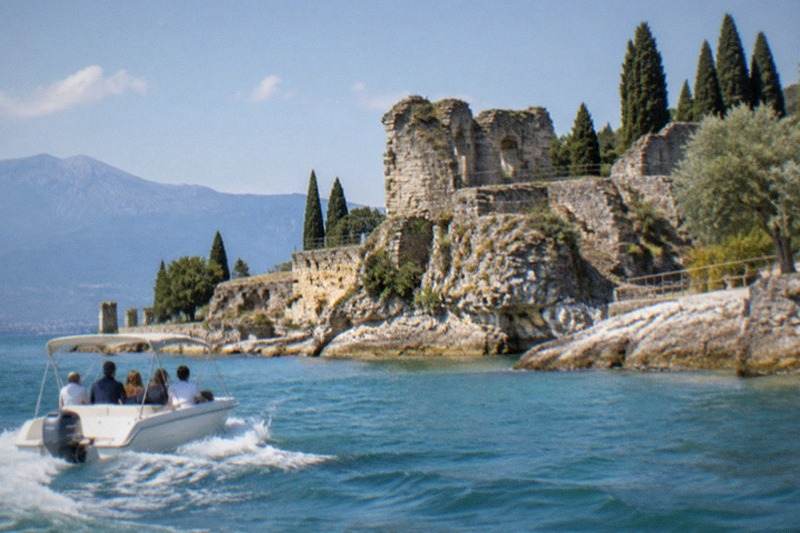A small open boat circles the tip of the Sirmione peninsula, its wake curling across turquoise shallows beneath the cliffs where the Roman ruins stand among olive trees.