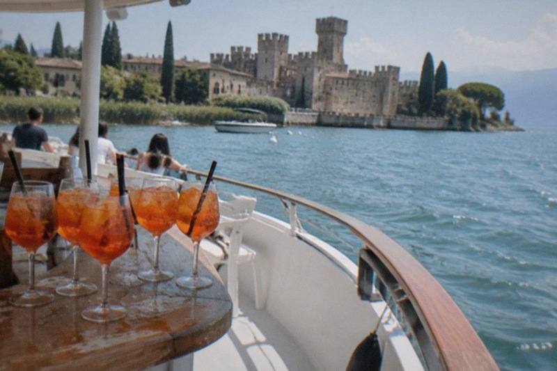 Orange spritz drinks sit on a wooden boat table as passengers look toward a Lake Garda lakeside stone castle across the water.