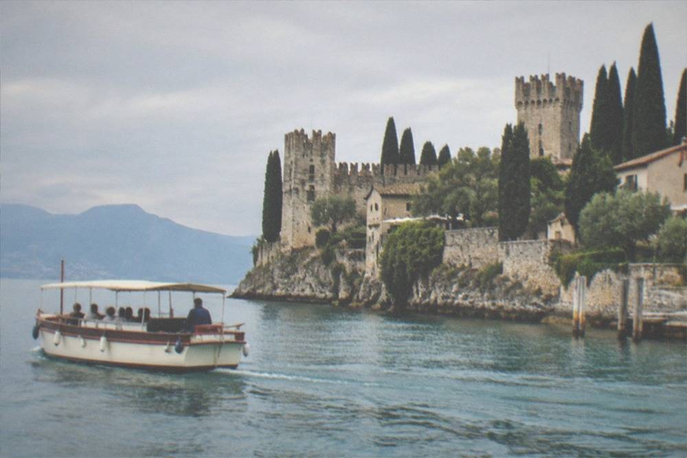 A small covered passenger boat with people aboard passes a rocky lakeshore lined with stone castle walls, towers and tall cypress trees, on Lake Garda.