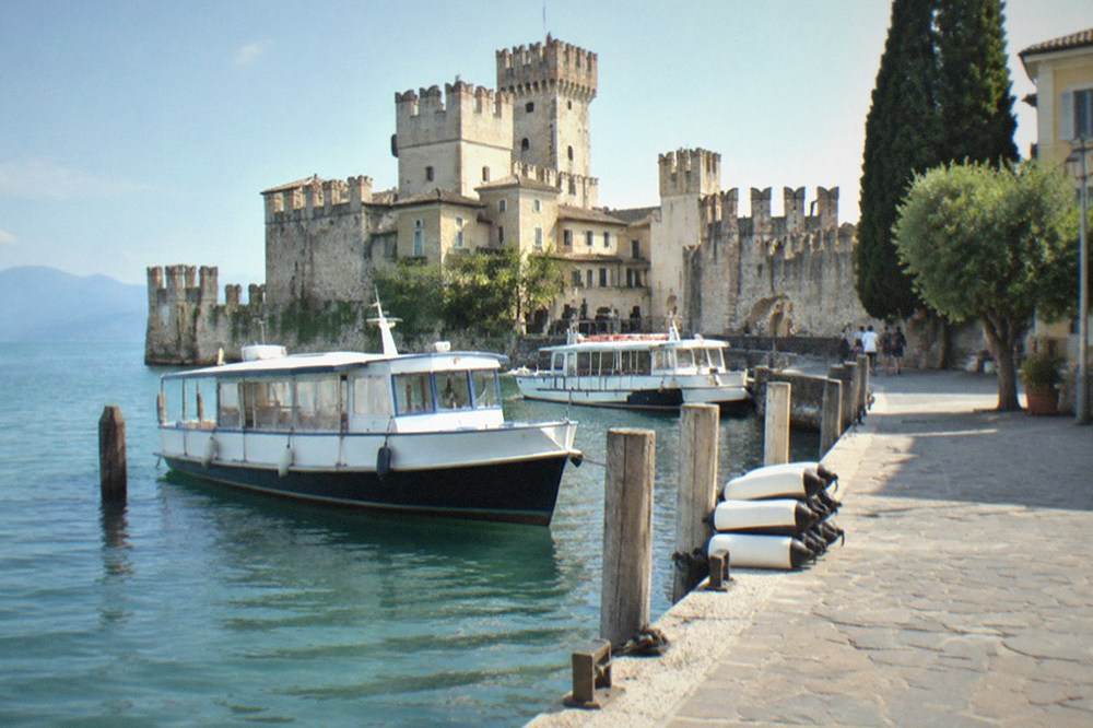 A small Lake Garda ferry landing stage with moored passenger boats and a lakeside promenade behind