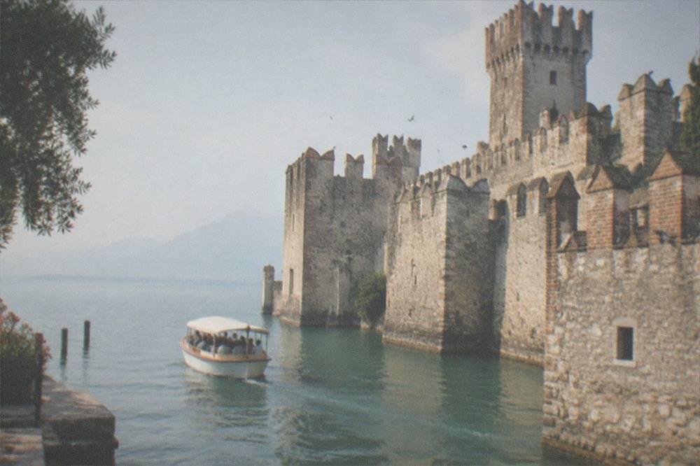 A small covered tour boat on Lake Garda beside a large stone castle with towers and battlements, hazy mountains beyond.