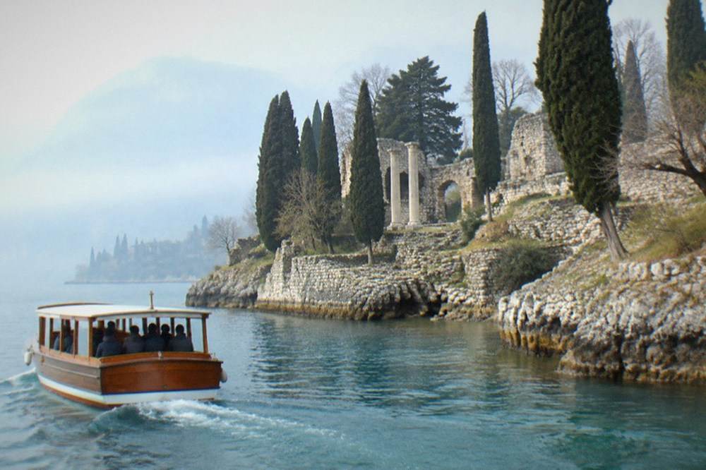 A wooden boat cruising past a rocky peninsula with cypress trees and a large Roman villa