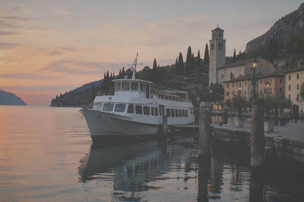 A white passenger boat moored at a dusk Lake Garda lakeside quay beside a stone church tower, cypress trees and distant mountains.