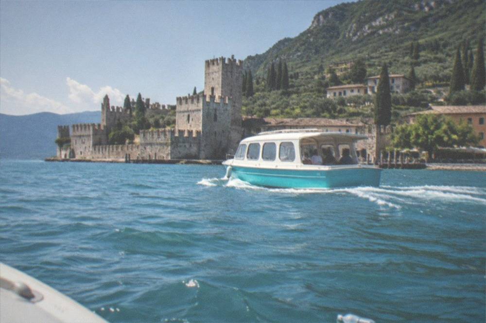 A small brightly coloured boat underway on open lake water with a distant castle town on the shoreline