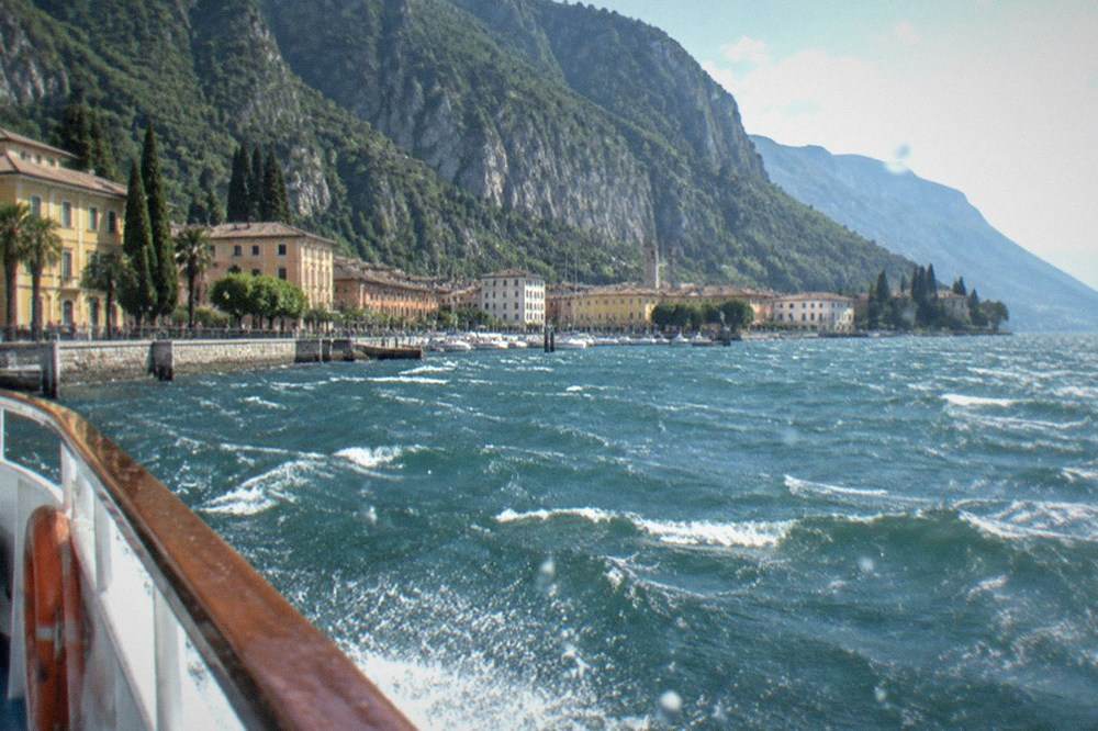 Choppy Lake Garda water from a boat railing, with a town of yellow buildings along the shore and a steep mountain rising behind it.