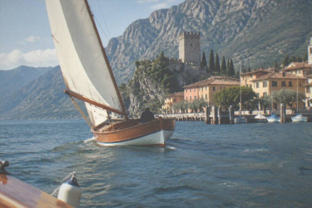 A wooden sailboat with white sails on Lake Garda beside a town, stone tower on a rocky hill, and mountains behind.