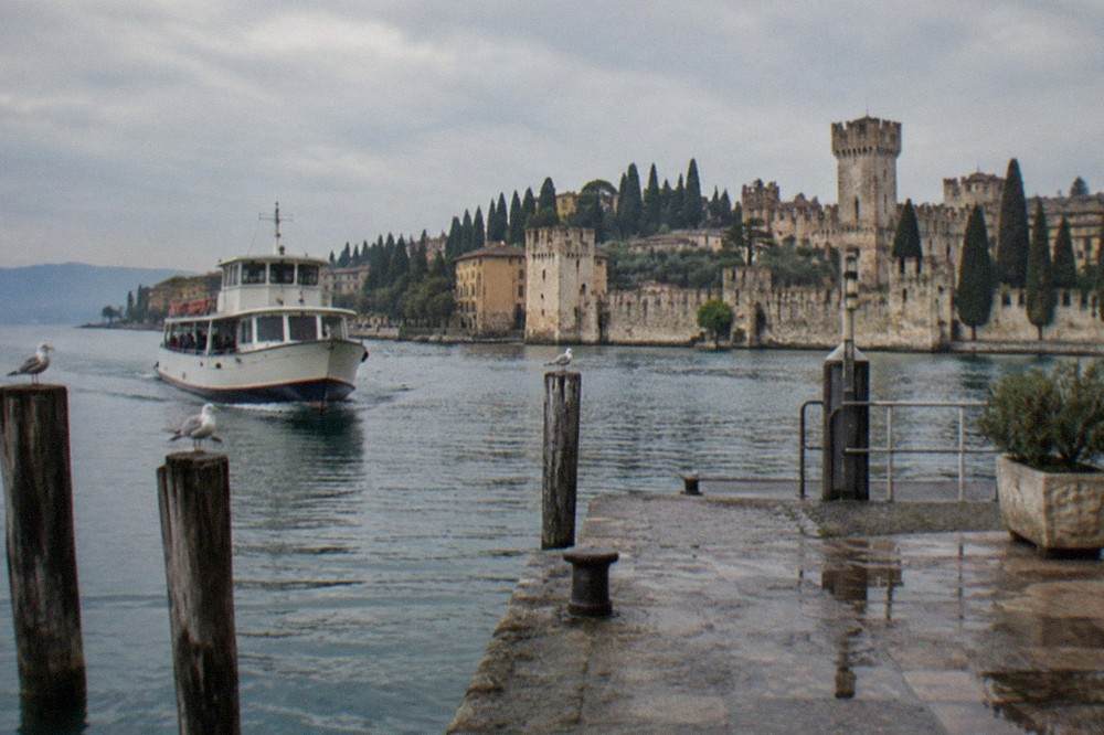 A small ferry approaching a stone landing stage with a walled old town and cypress trees above the water