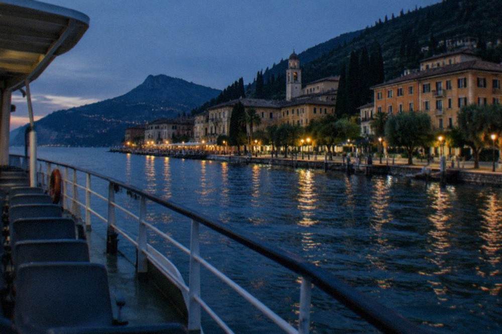 A ferry passenger deck viewed across the water with a walled town and distant mountains on the shoreline