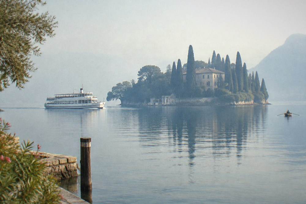 A white passenger ferry on a misty lake passes a small island of tall cypress trees and a villa, with a stone pier in the foreground, on Lake Garda.