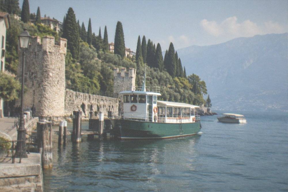 Small covered boat and larger white ferry on Lake Garda beside a hillside castle, cypress trees and lakeside buildings, mountains in the distance.