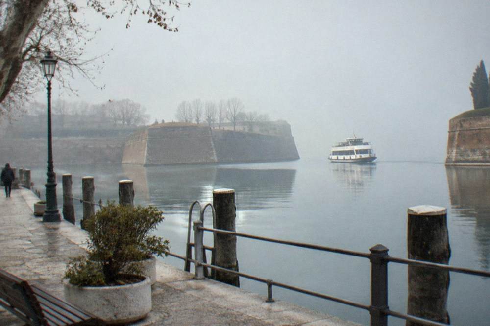 Foggy stone promenade with lamp, planter and posts beside calm water, a person walking, and a white boat between low fortress walls, on Lake Garda.