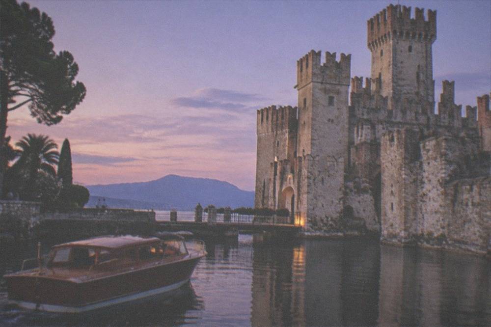 A wooden motorboat sits in still water beside a stone castle with towers at dusk, trees at left and mountains beyond, on Lake Garda.