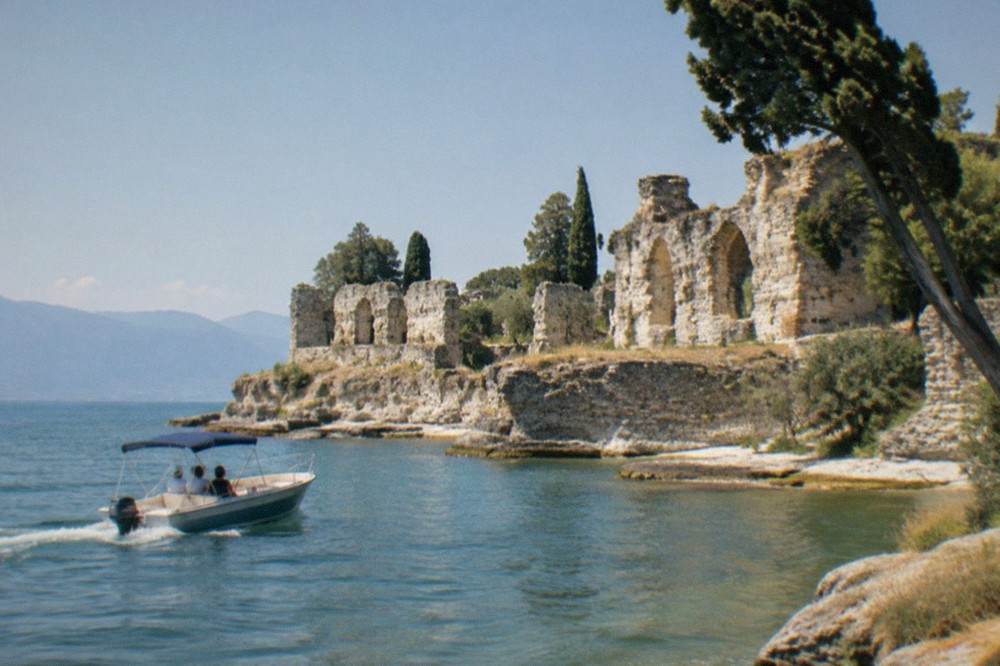 A canopied motorboat with passengers passes crumbling stone ruins on a rocky lakeshore, trees and distant mountains beyond, on Lake Garda.
