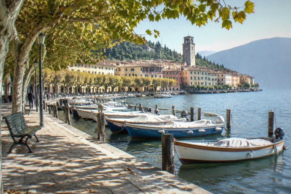 A lakeside promenade with parked boats on the water and a stone tower in the background