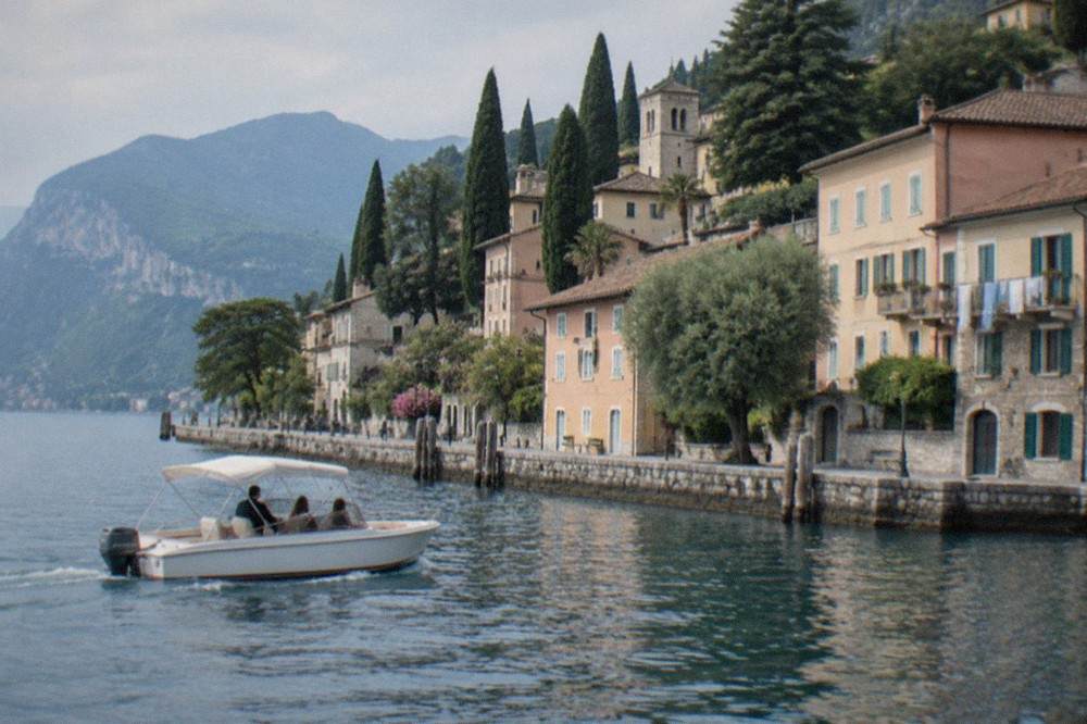 A low open passenger boat gliding along a shoreline of pale stone buildings and cypress trees on Lake Garda