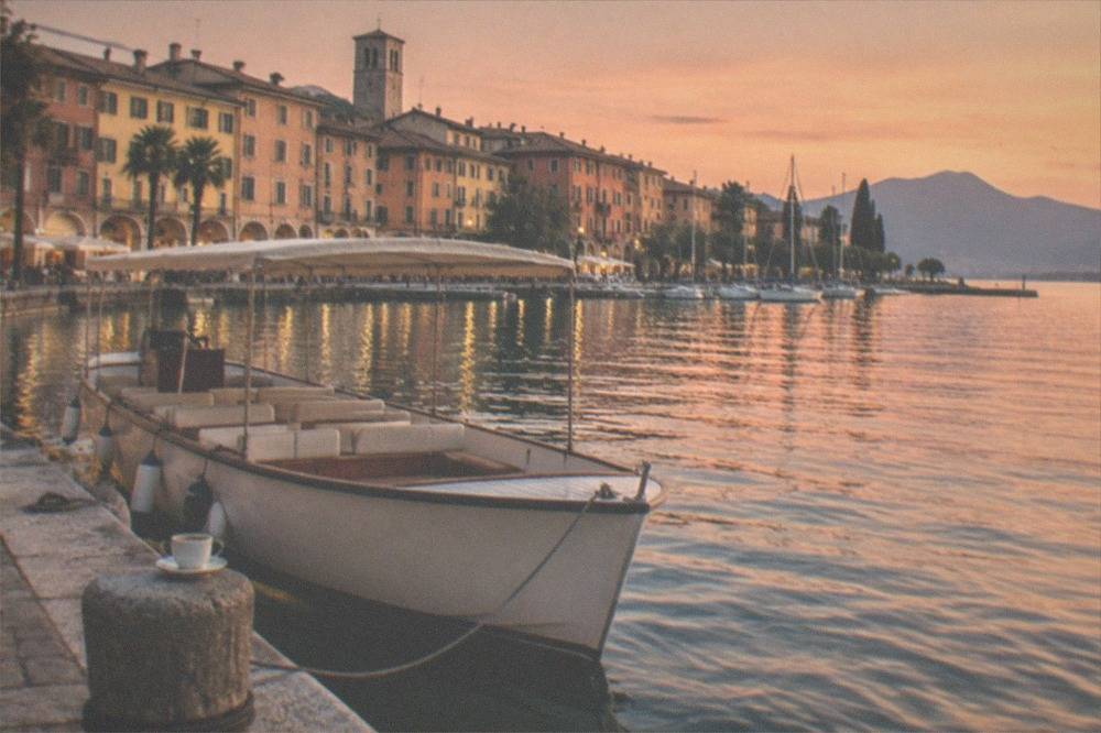 An open tour boat moored at a stone jetty in front of pastel coloured buildings on a Lake Garda evening