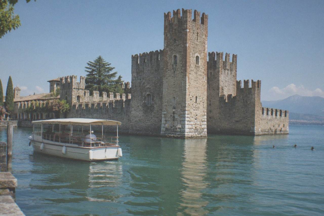 A small passenger boat cruising past a stone castle tower rising from the water at Sirmione on Lake Garda