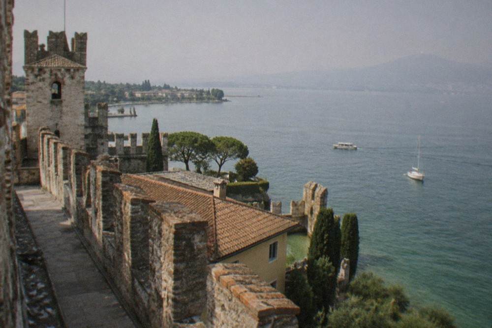 Crenellated stone walls of a lakeside fortress with a view over red rooftops and open water beyond.