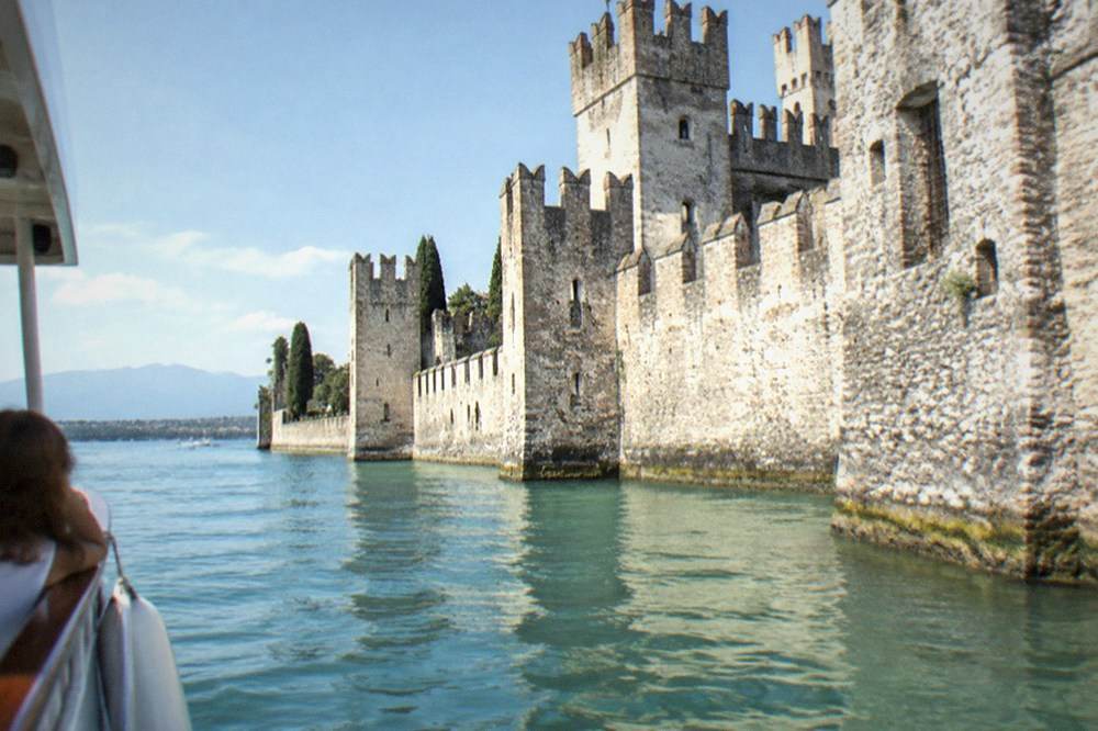 A small passenger boat passing close to stone fortress walls that rise directly from the water of a lake harbour.