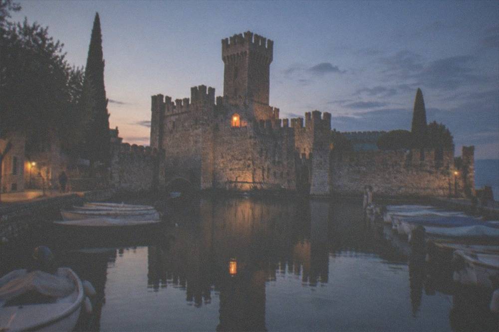 A harbour at dusk with a stone fortress tower reflected in still water alongside rows of moored small boats.