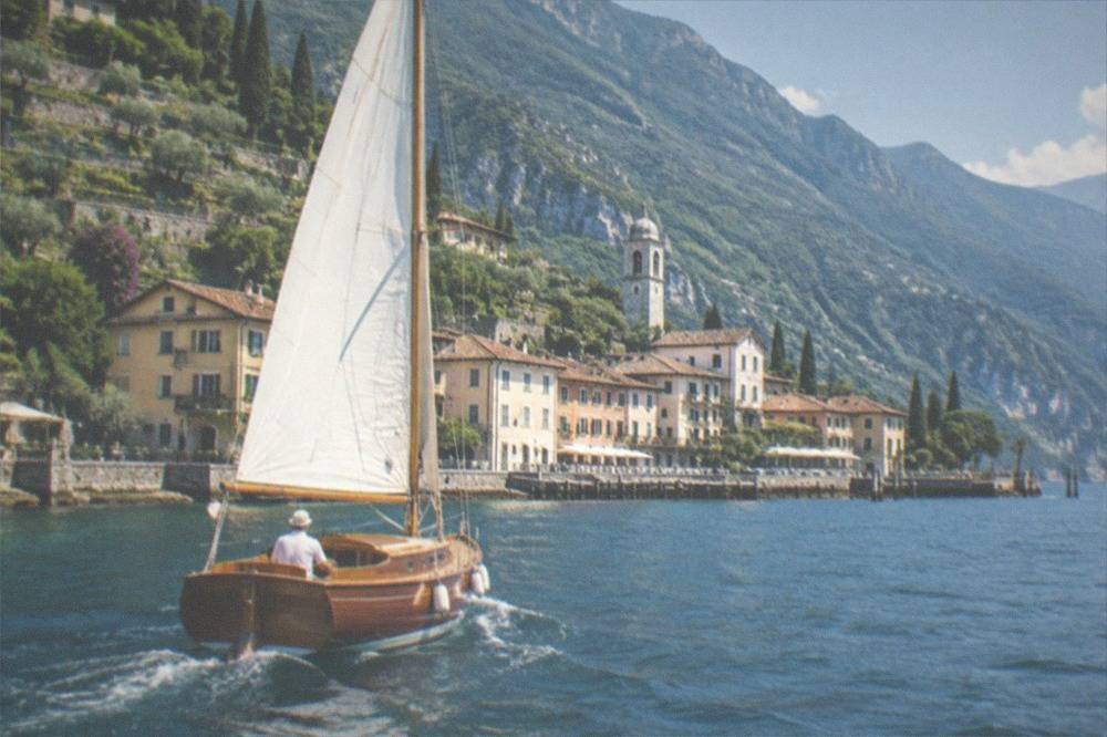 A wooden sailboat cruising on the northern part of Lake Garda with Riva del Garda's rooftops and mountains in the distance.