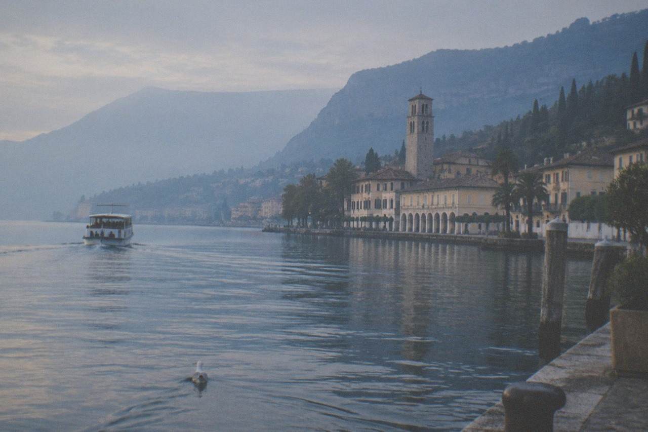 A small passenger boat crossing calm water toward a lakeside town with a stone tower