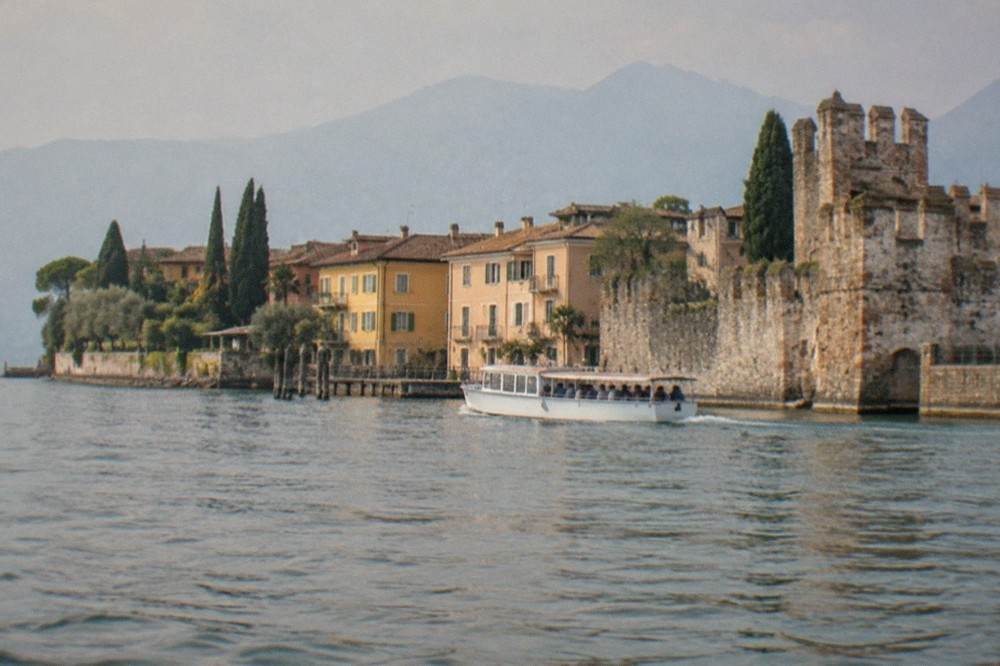 A small passenger boat cruising close to the Sirmione peninsula shoreline on flat water