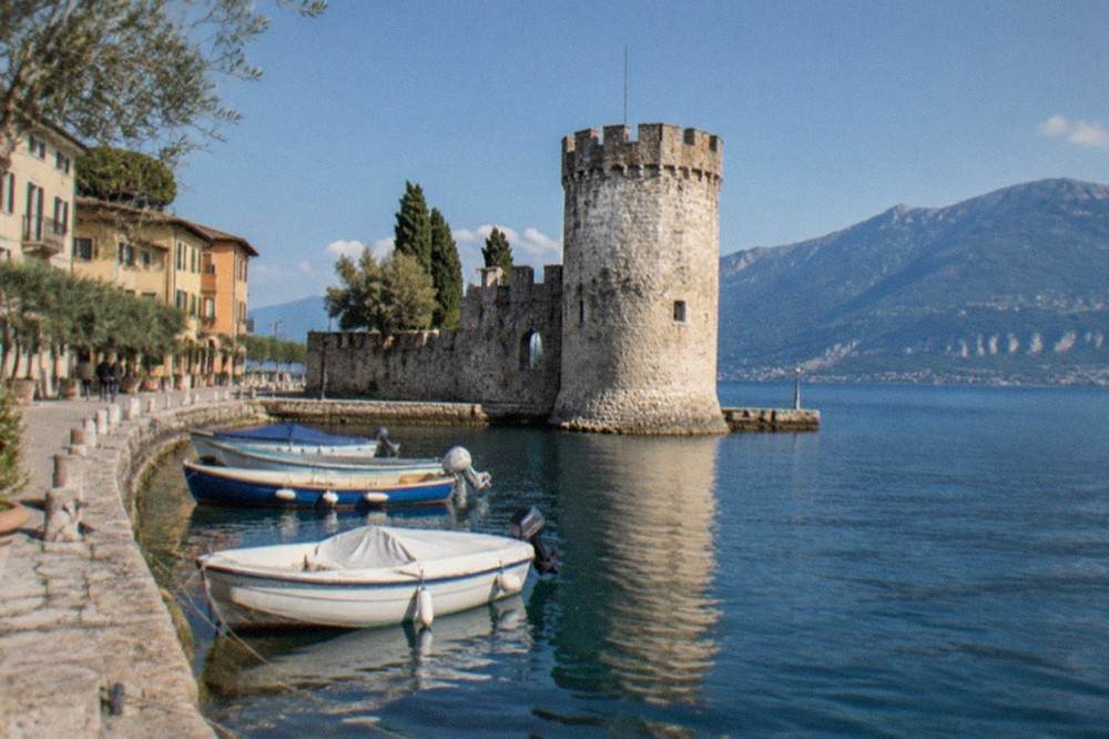 A stone castle tower at the water's edge with small boats moored along a lakeside promenade