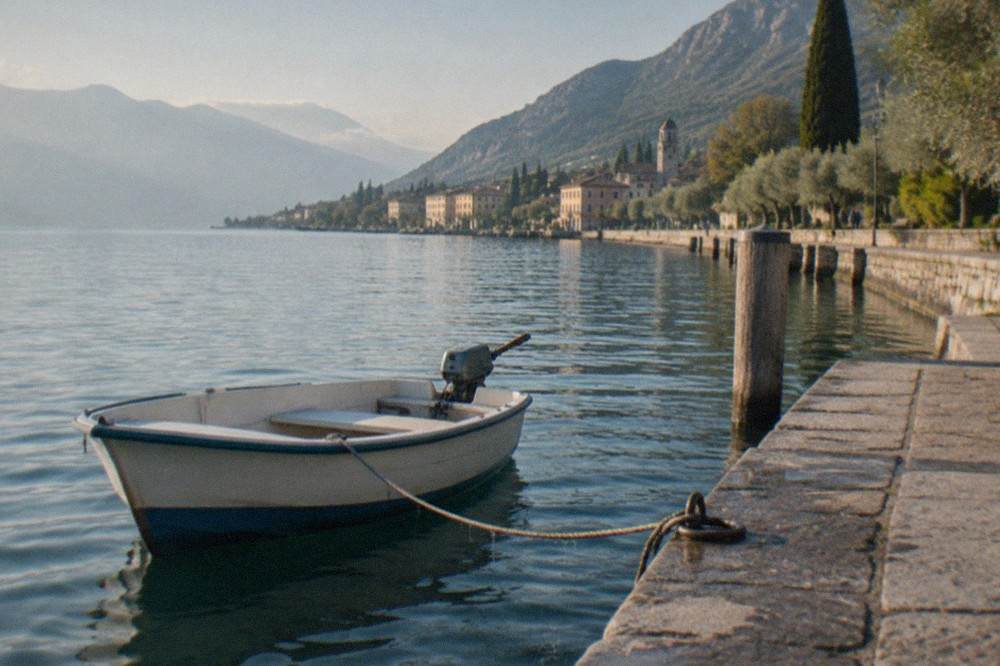 A small white motorboat with a blue hull is tied by rope to a stone quay beside a Lake Garda lakeside town and mountains.