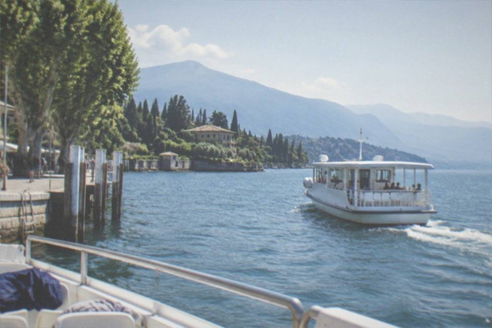A wide passenger boat cruising on Lake Garda in afternoon sun with mountains on the far shore and jackets folded on the seats