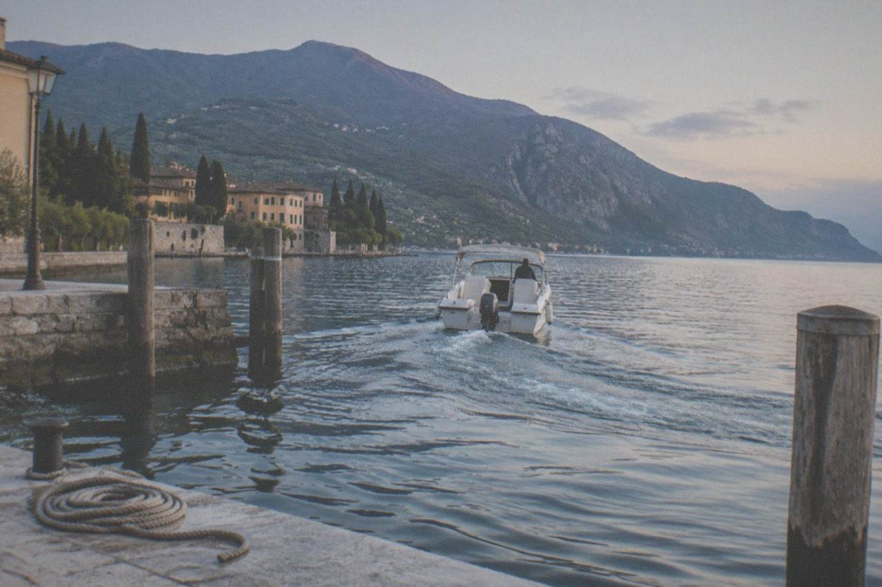 A calm Lake Garda at morning with a small boat leaving a stone harbour and mountains rising behind the water