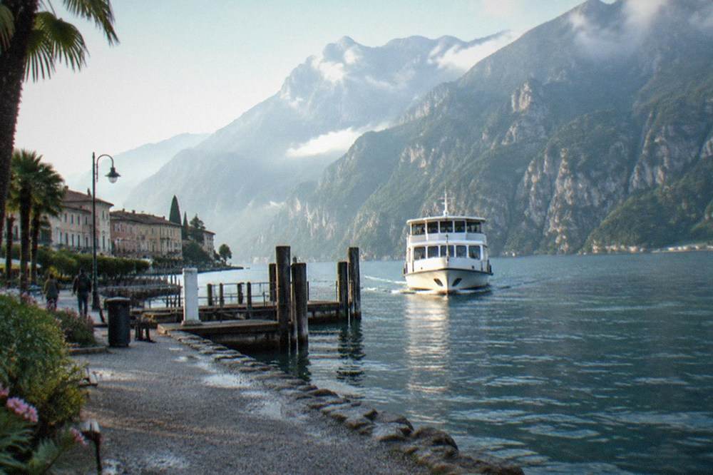 White passenger boat nears wooden dock posts on a cobbled Lake Garda lakeside promenade, with mountains and buildings beyond.