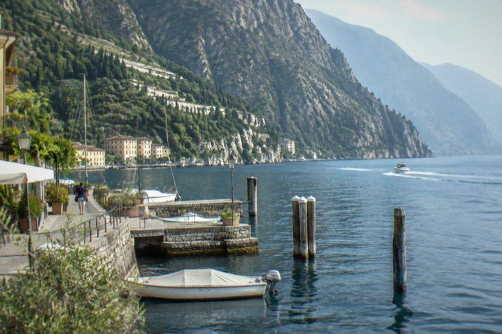 Stone Lake Garda lakeside walk with a covered motorboat, wooden posts and a pedestrian, yellow buildings and steep forested mountains across blue water.