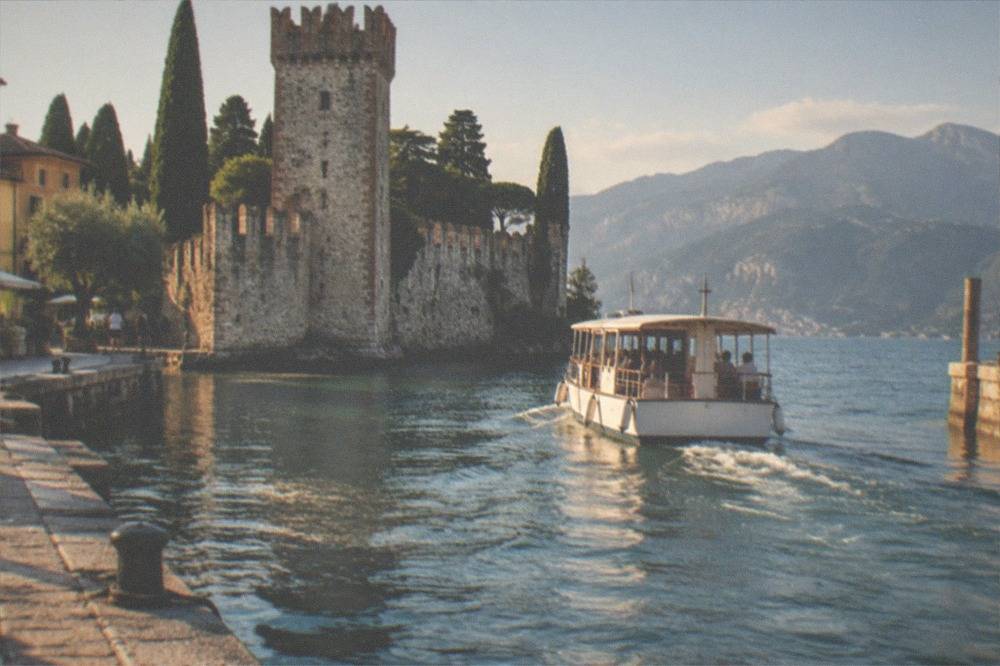 A passenger boat leaving a small harbour with a stone castle tower and cypress trees in the background.