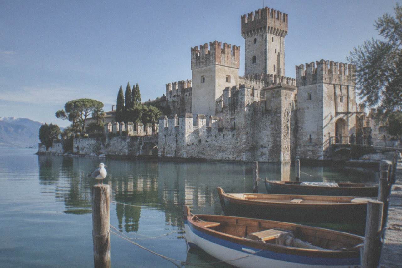 Wooden boats moored in clear water below a medieval stone castle on a peninsula reaching into Lake Garda.
